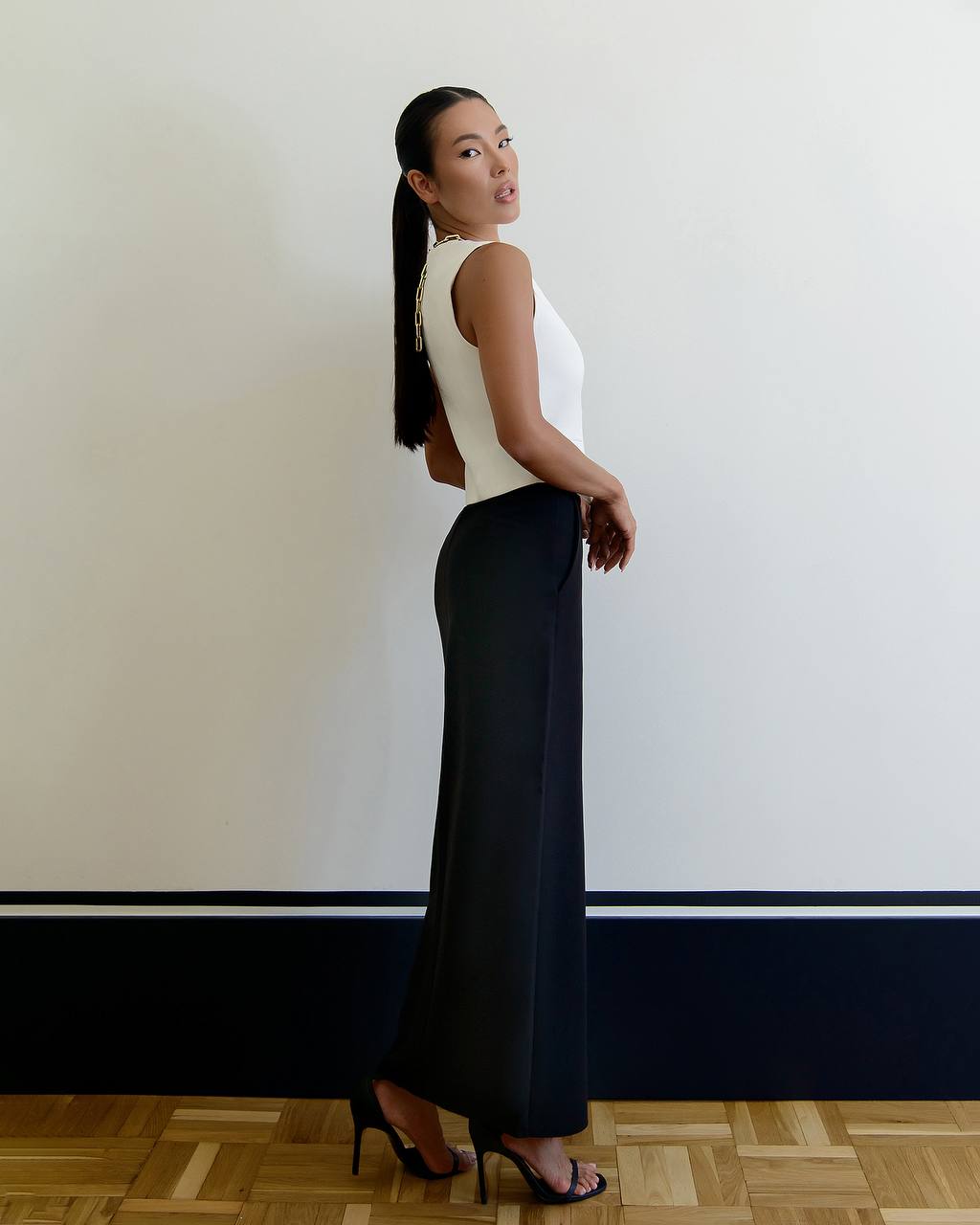 Woman in a white sleeveless top and black wide-leg pants standing against a plain wall.