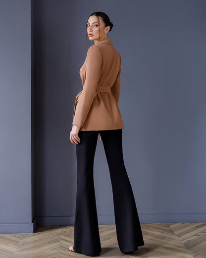 Woman wearing a tan top and black pants against a gray wall.
