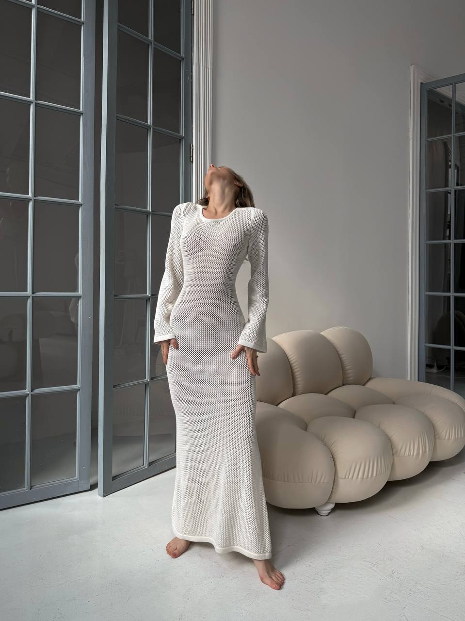 Woman in a long white dress standing in a modern room with a unique sofa.