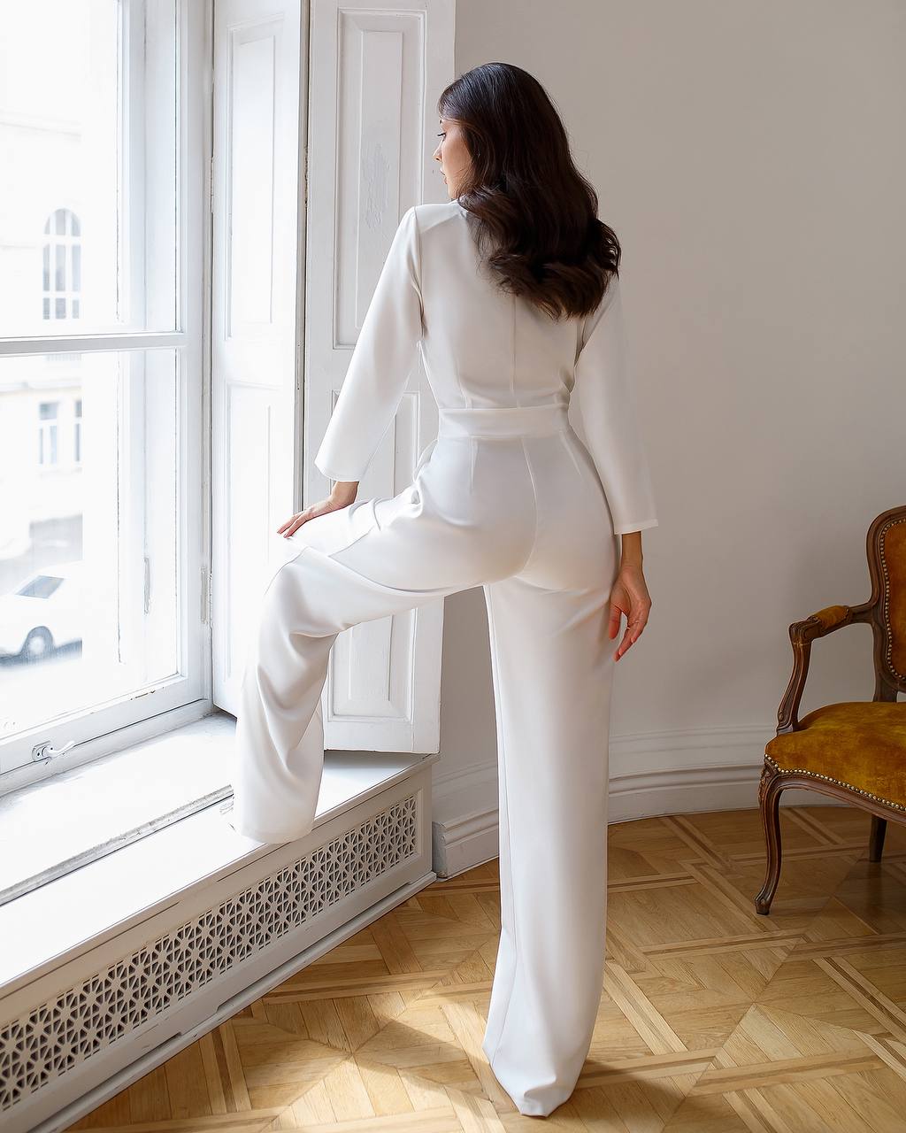 Woman in a white outfit standing by a window in a room with wooden flooring.
