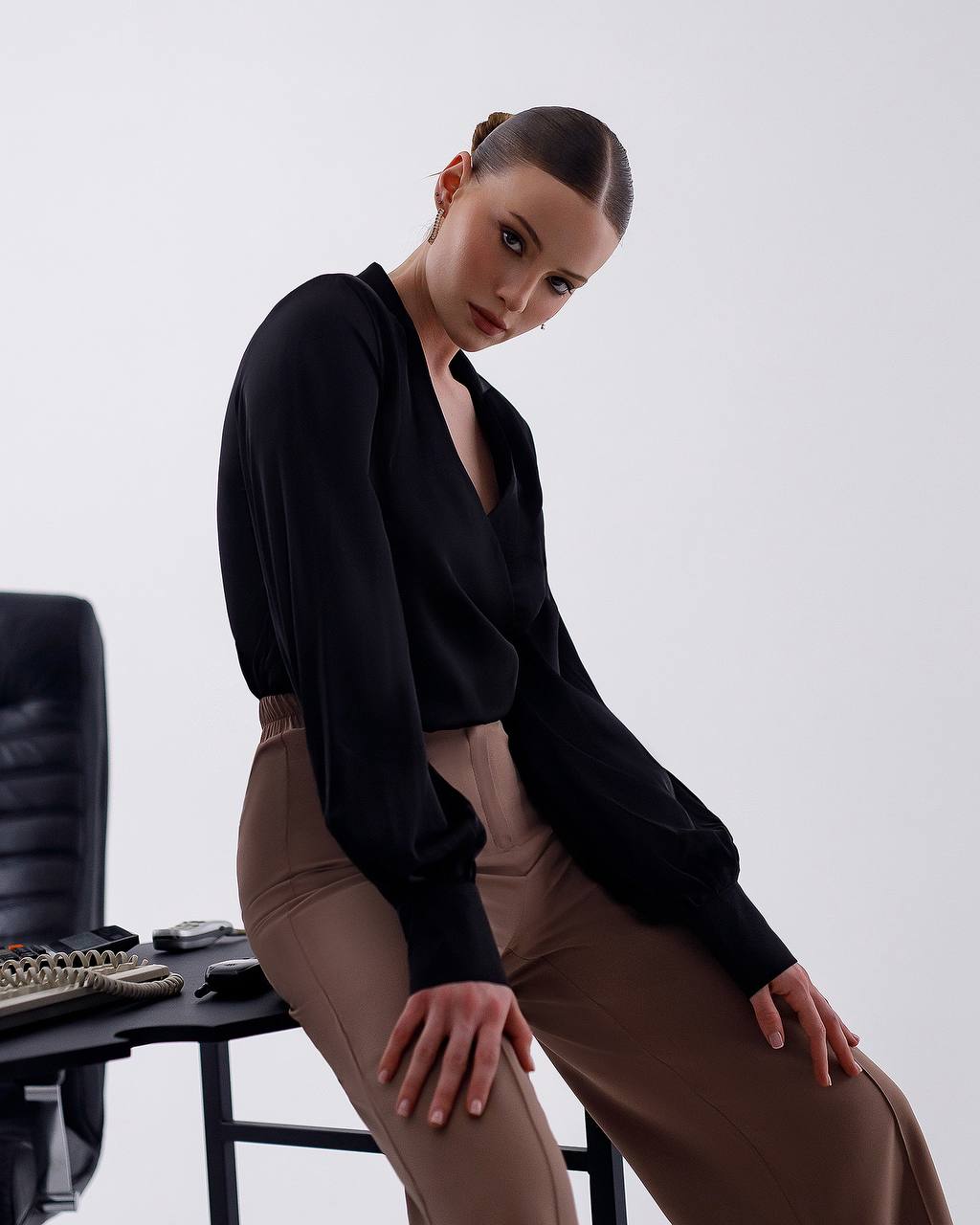 Woman in black top and brown pants sitting on a chair against a white background