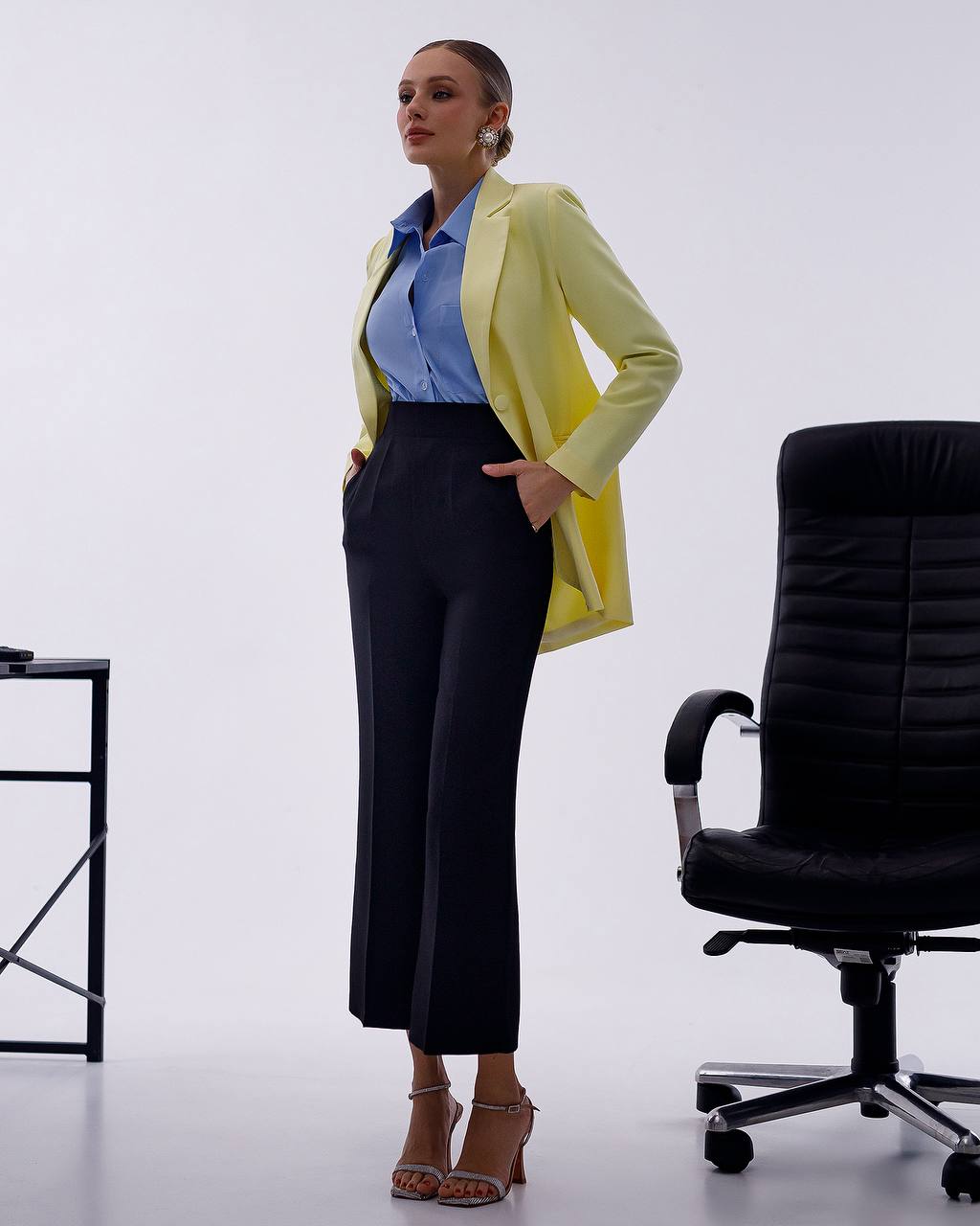 A woman models a light yellow blazer over a blue button-down shirt and black wide-leg cropped trousers, standing in a studio next to a black office chair.