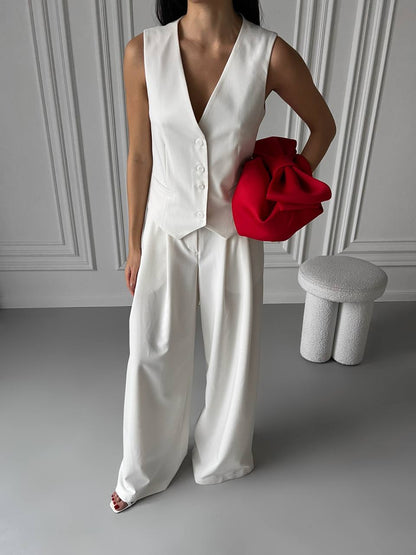 Person wearing a white sleeveless top and pants holding a red bag in a minimalistic setting.
