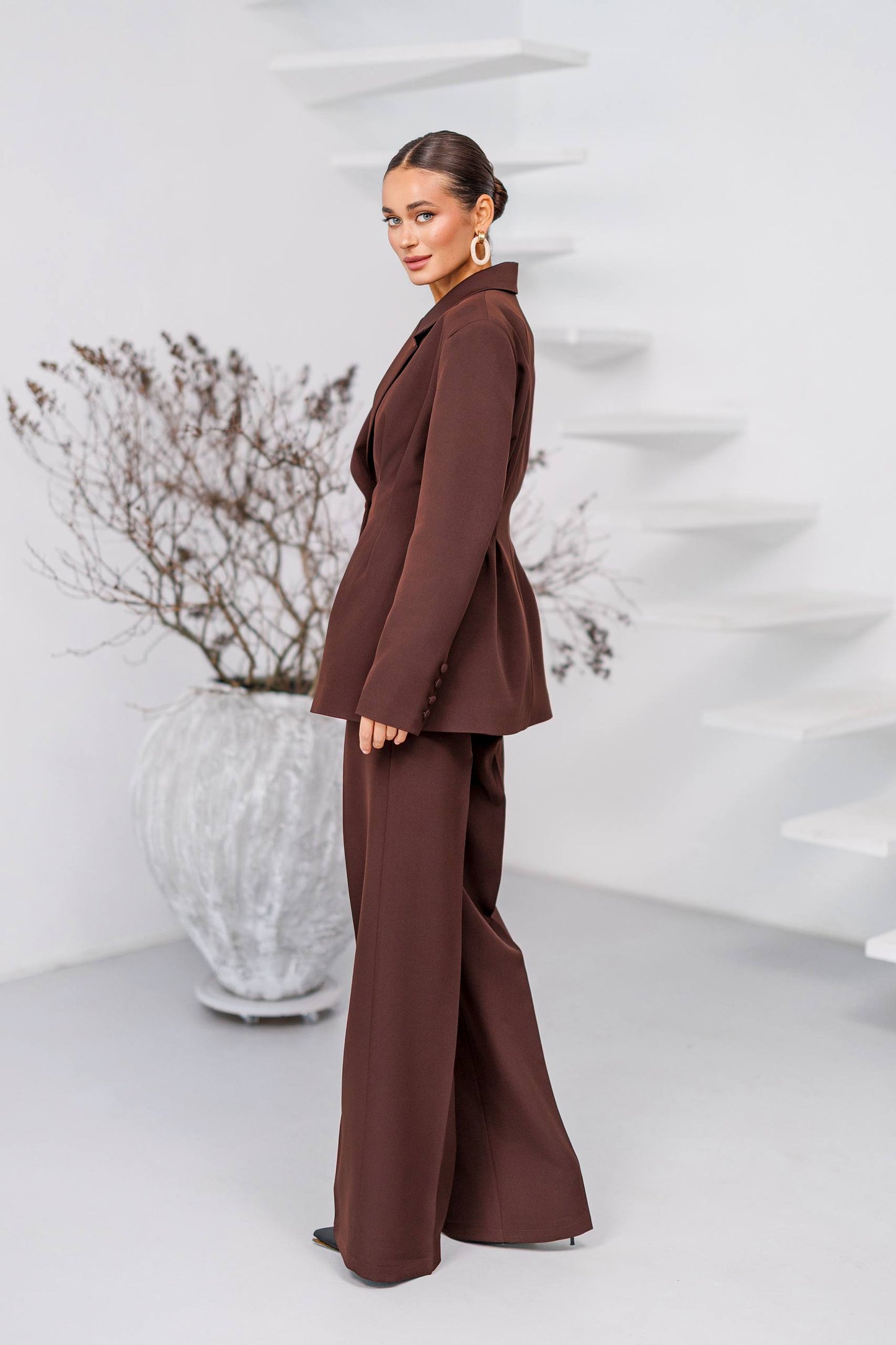 Woman in a brown suit standing in a minimalistic room with a plant.