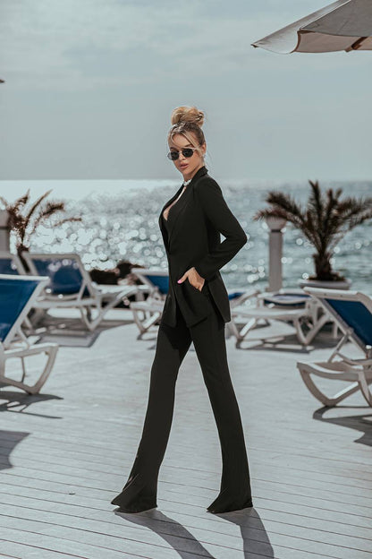 Woman in a black suit standing on a beach with lounge chairs and umbrellas in the background.