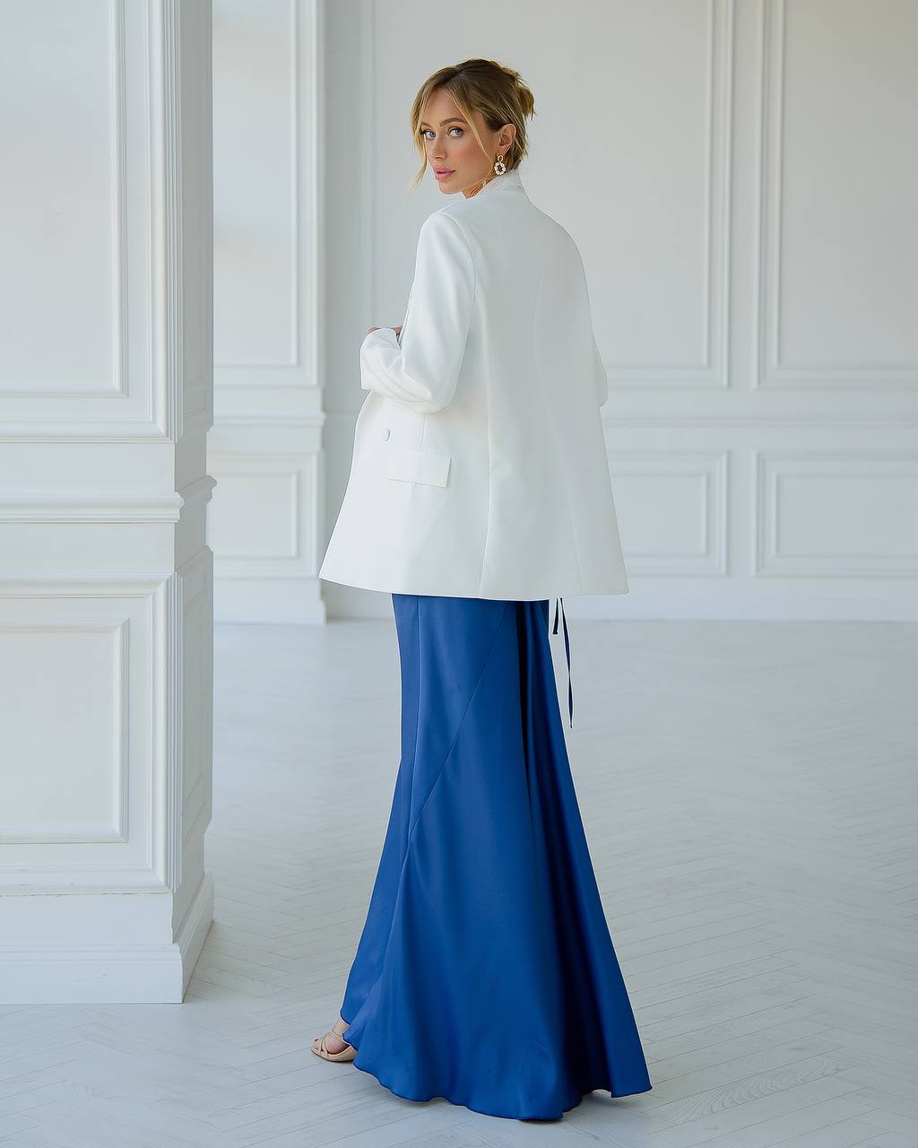 Woman wearing a white coat over a blue dress in a room with white walls.
