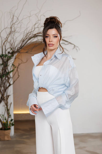 Woman wearing a light blue shirt over a white top with white pants, standing indoors.