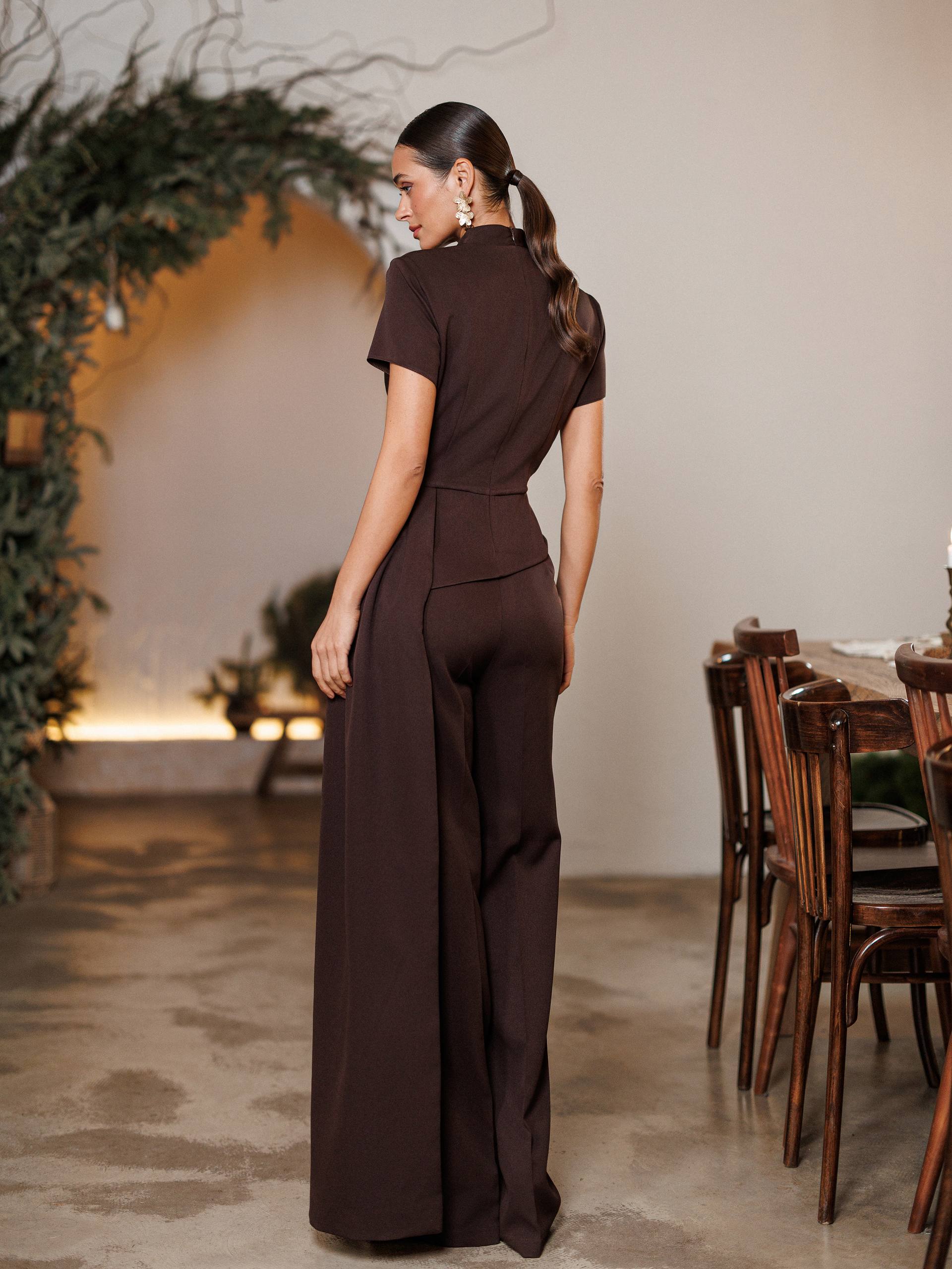 Woman in a brown jumpsuit standing in a room with wooden furniture and decorative plants.
