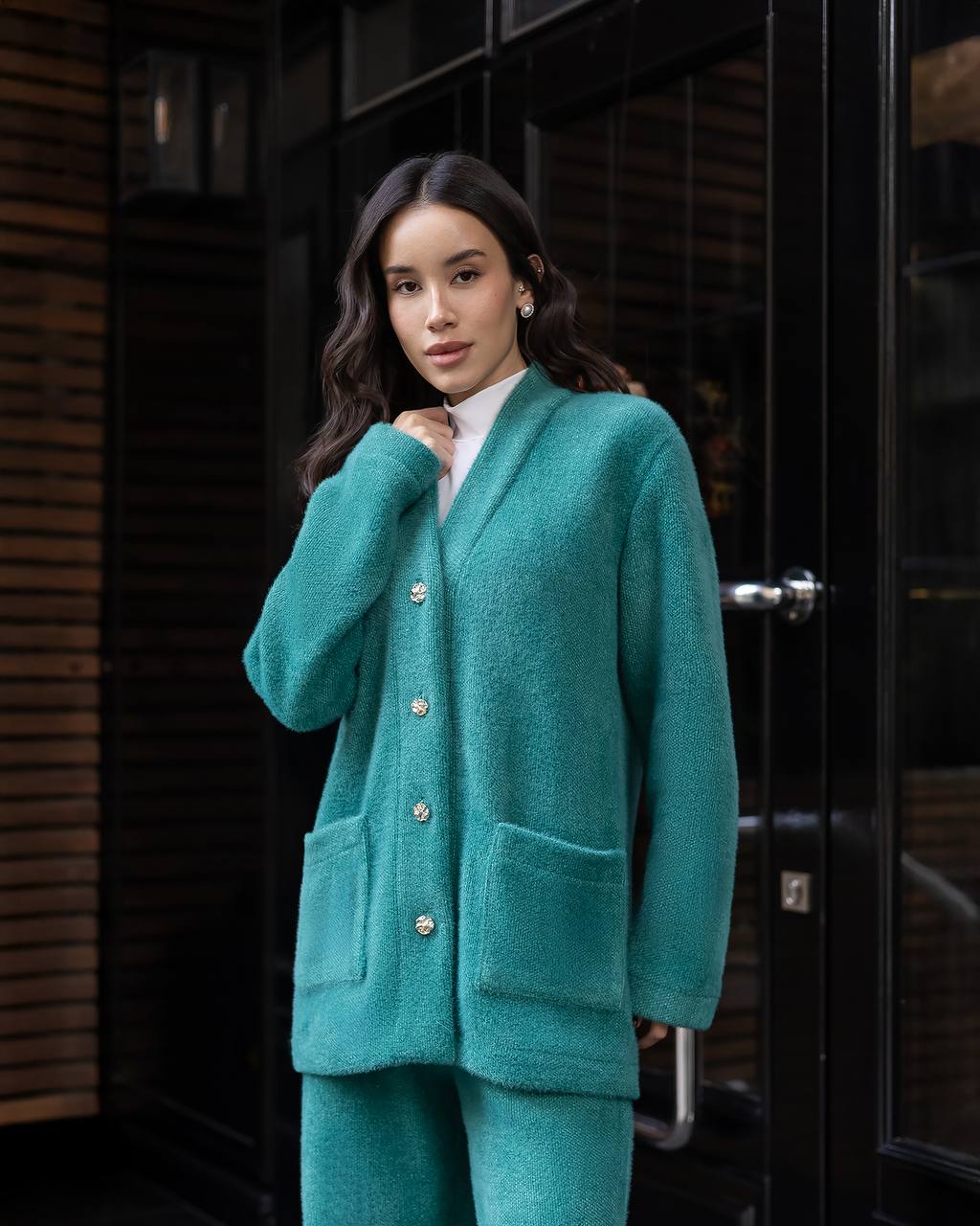 Woman wearing a teal cardigan and pants standing in front of a dark door.