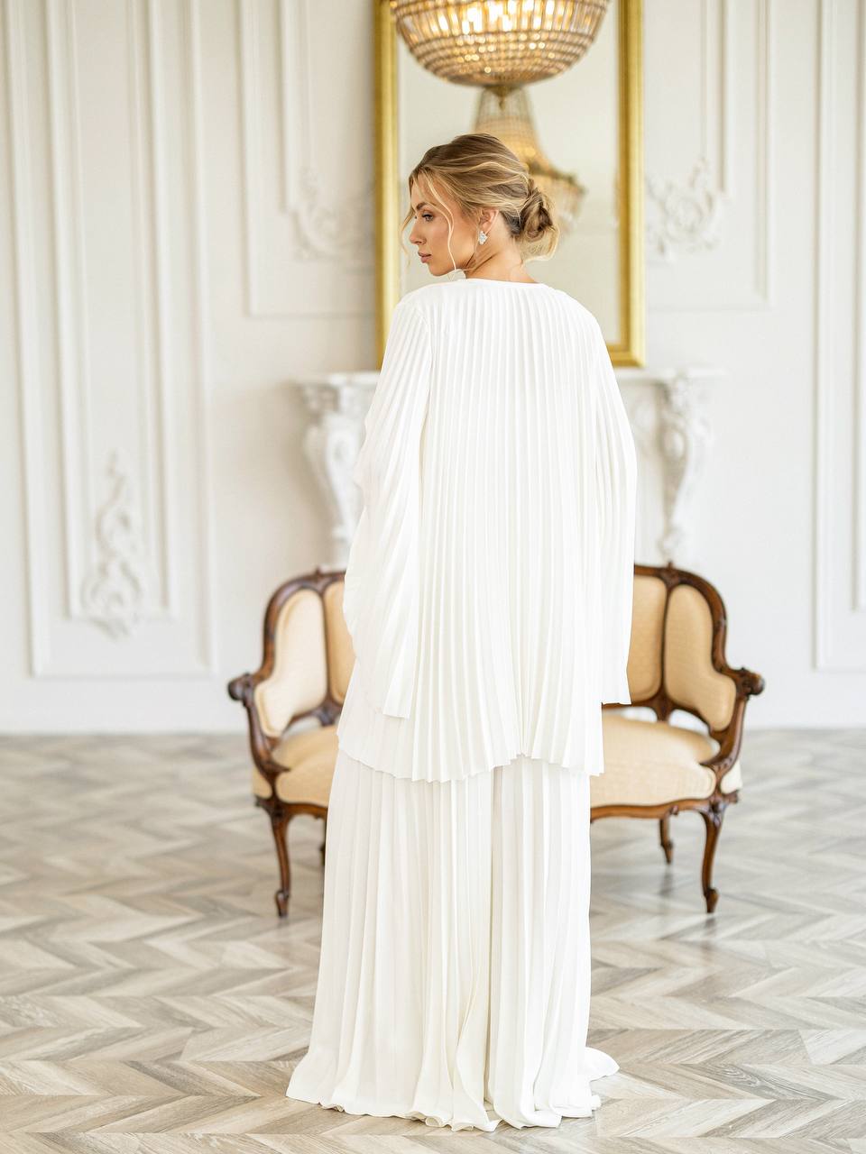 A woman stands in a bright, elegant room wearing a flowing white pleated dress with a draped cape-style overlay. The room features classic white paneled walls, a gold-framed mirror, a white console table, and two vintage-style armchairs with beige upholstery and dark wood frames.