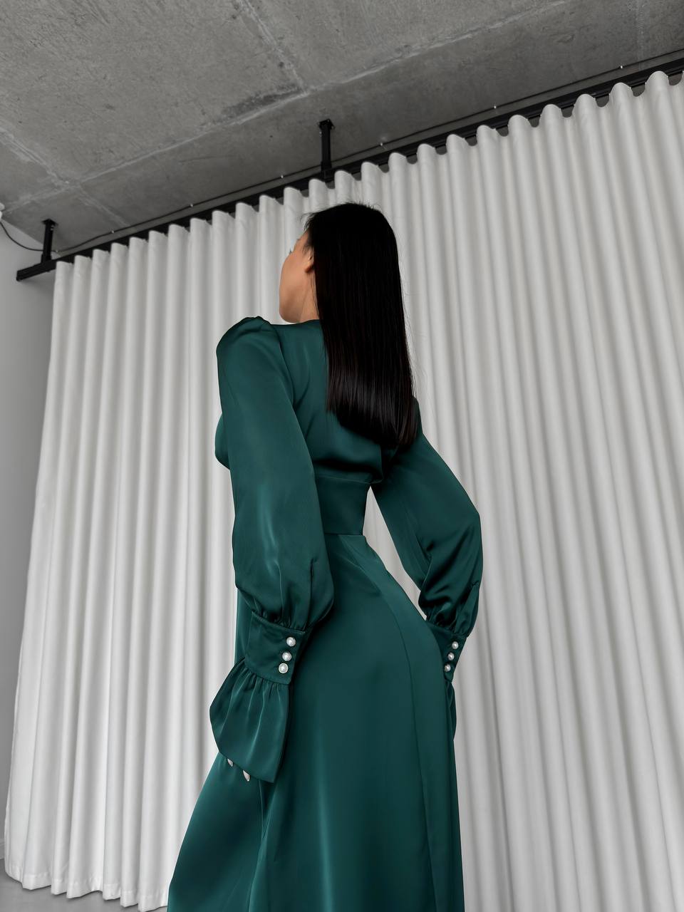 Woman in a green dress standing in front of white pleated curtains.