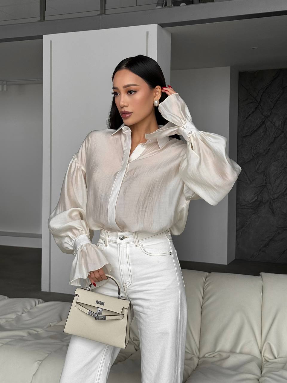 Woman wearing a sheer white blouse with puffed sleeves and white pants, holding a beige handbag in a modern interior setting.