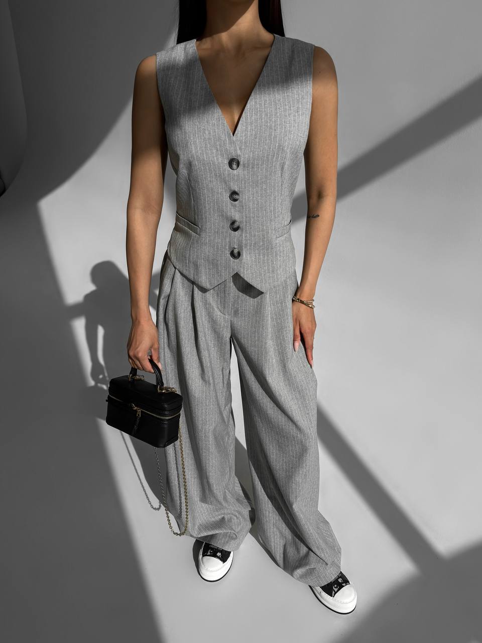 Person wearing a gray vest and wide-leg pants holding a black handbag on a plain background