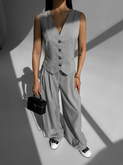 Person wearing a gray vest and wide-leg pants holding a black handbag on a plain background