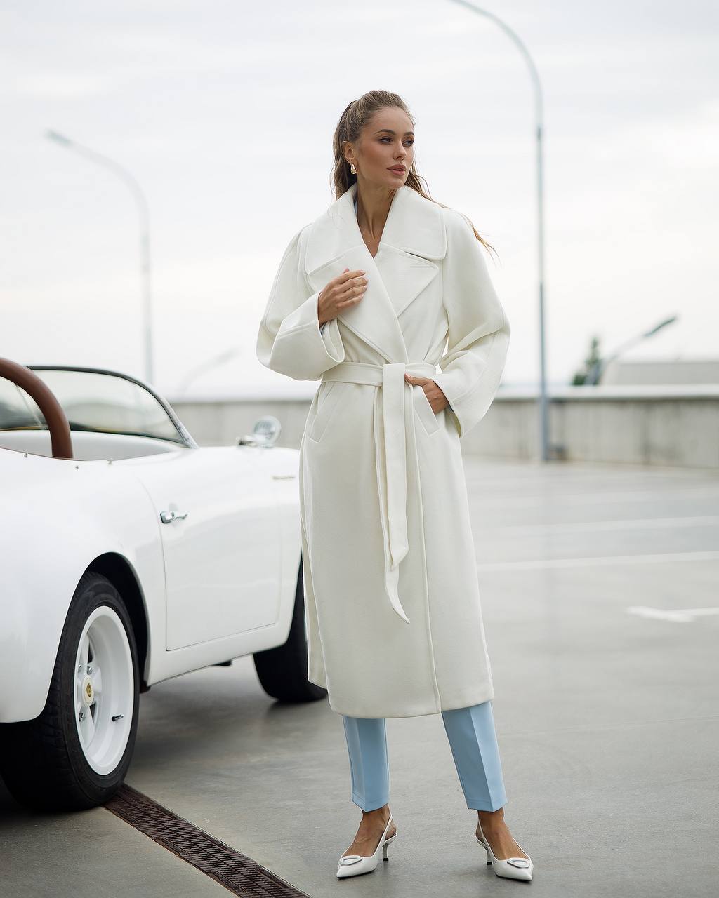 Woman in a white coat standing next to a white car on a road.