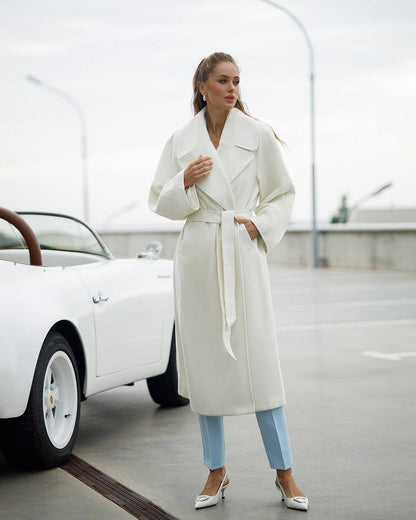 Woman in a white coat standing next to a white car on a road.
