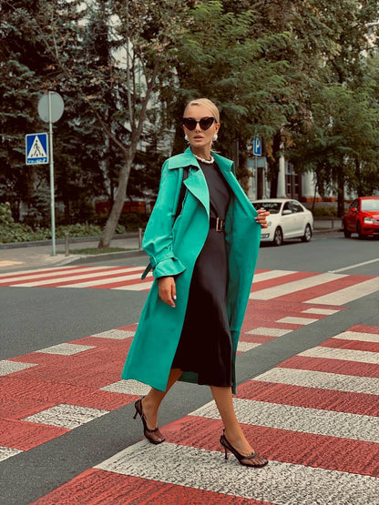 Woman in a teal coat crossing a street with trees and cars in the background
