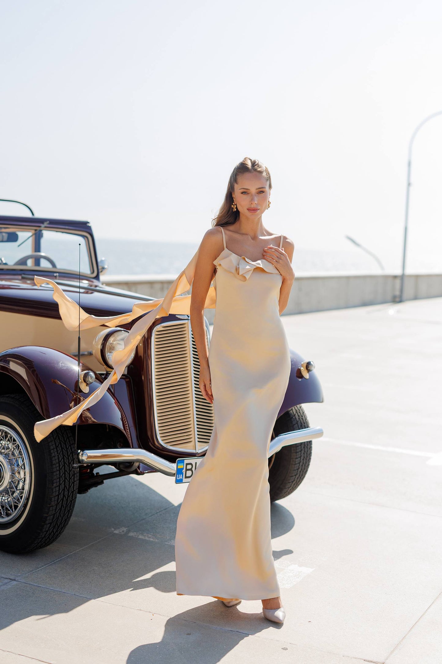 Woman in a long, elegant dress standing next to an old-fashioned car on a sunny day.