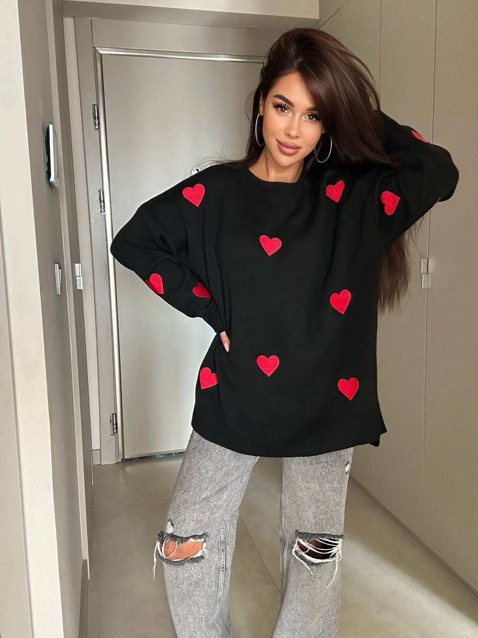Woman wearing a black sweater with red heart patterns and gray ripped jeans indoors.
