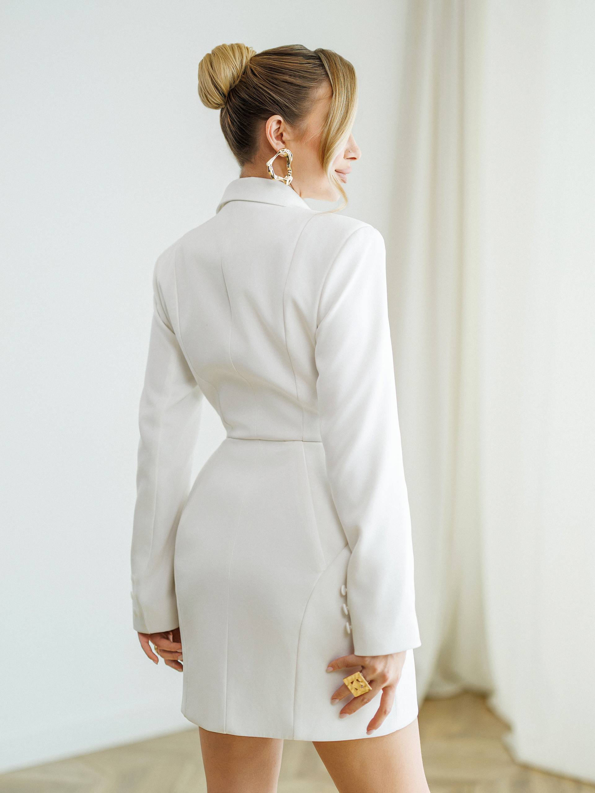 A woman wearing a white long-sleeve mini blazer dress, shown from the back in a bright indoor setting with white curtains and a light wood floor.