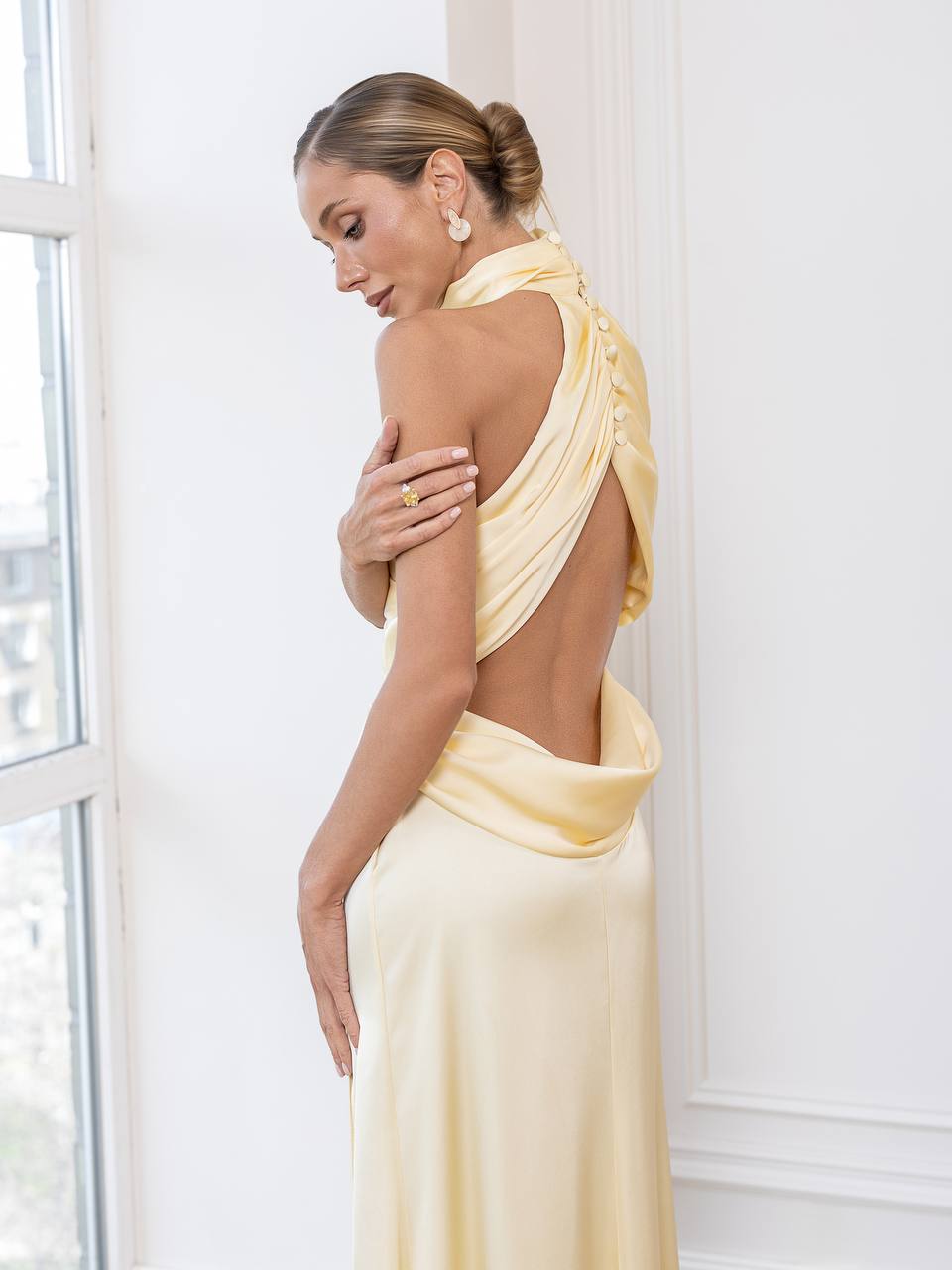 A woman wearing a pale yellow draped halter-neck gown with an open back, standing in a bright white room near a window.