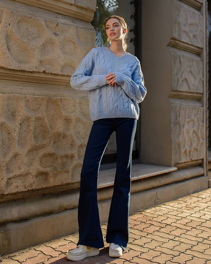 Person wearing a light blue cable knit sweater and dark blue flared jeans standing in front of a stone building.