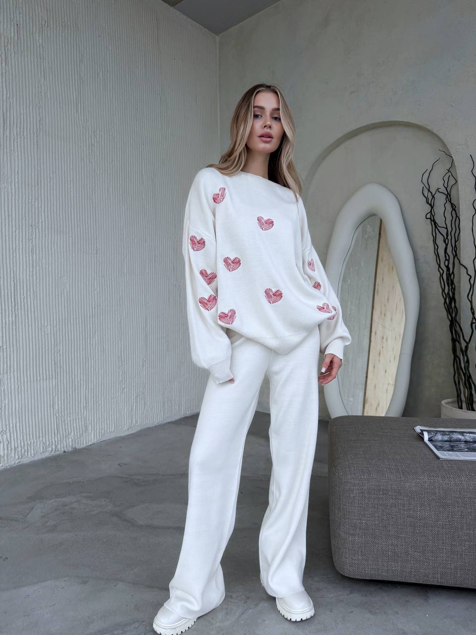 Woman wearing a white sweater with pink heart patterns and white pants in a minimalistic room.