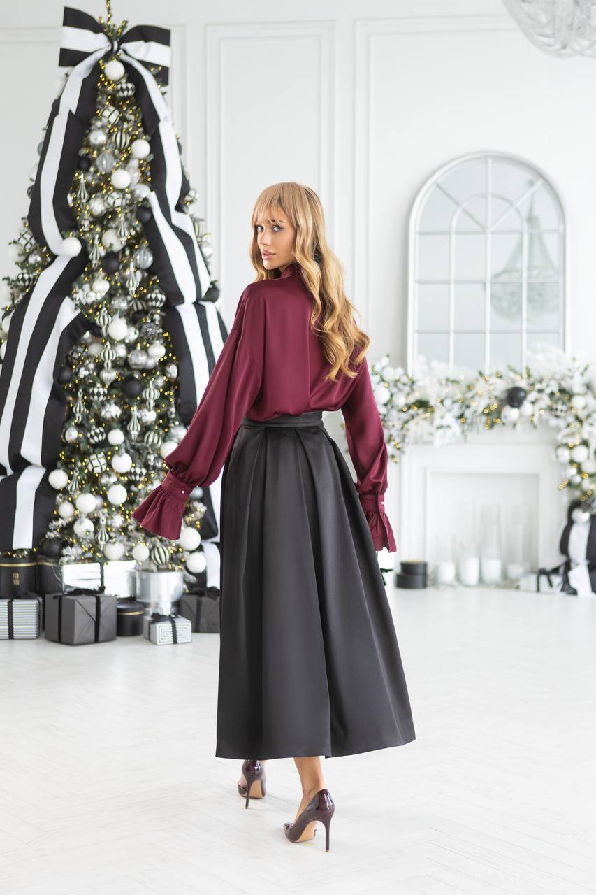 Woman in a burgundy blouse and black skirt standing in a decorated room with Christmas trees and presents.