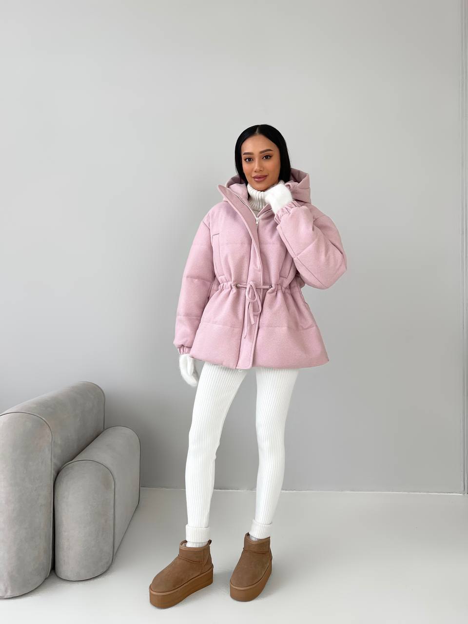 Person wearing a pink puffer jacket with a hood, white pants, and brown boots in a minimalistic setting.