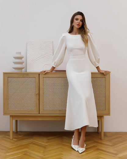 Woman in a white dress standing next to a wooden cabinet with a neutral background