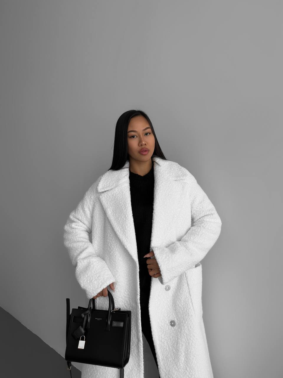 Woman wearing a white coat holding a black handbag against a gray background