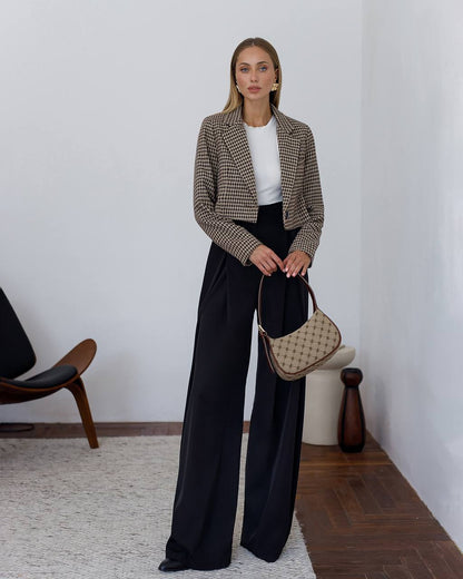 Woman in a stylish outfit with a jacket, white top, and black pants, holding a handbag in a minimalistic room.