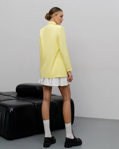 A model is shown from the back wearing a pale yellow blazer over a white pleated mini skirt, styled with white socks and black loafers, standing next to a black leather bench against a light gray studio background.