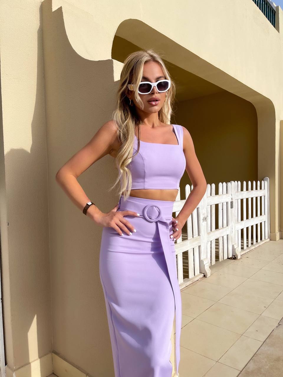 Woman in a lavender outfit standing against a beige wall with a white picket fence in the background.