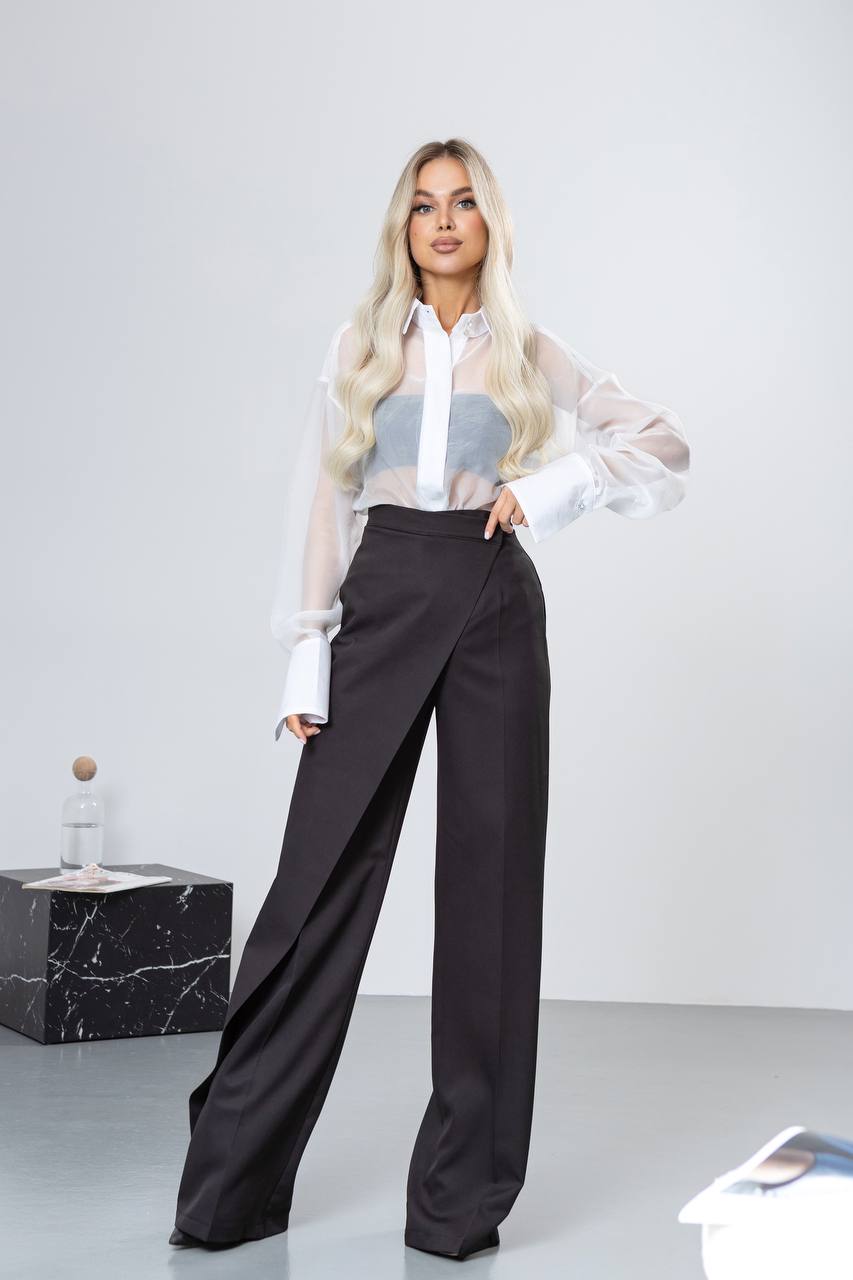 Woman wearing a white blouse with sheer sleeves and black pants on a plain background