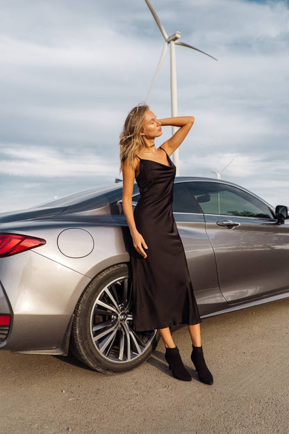 Woman in a black dress standing next to a silver car with a wind turbine in the background