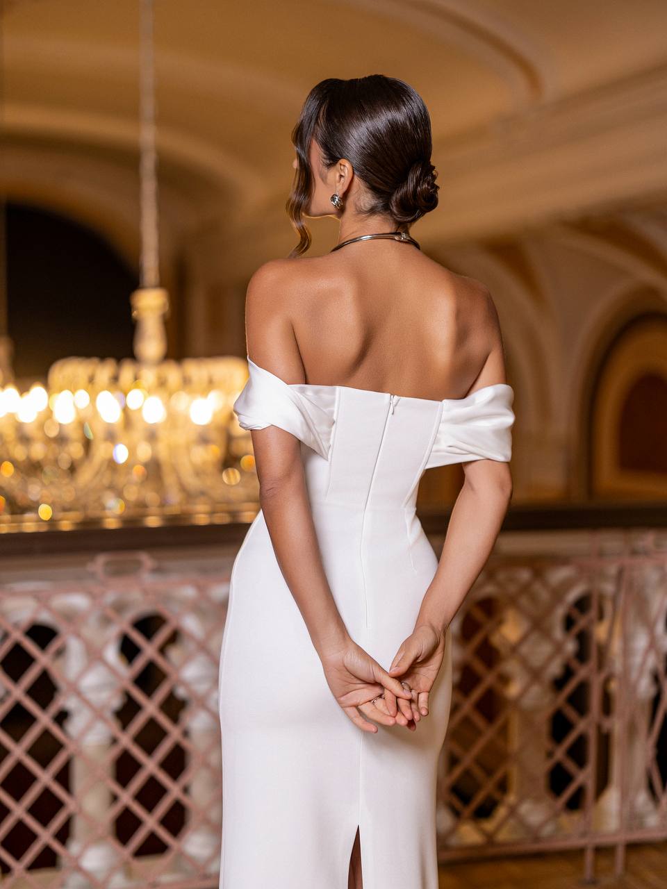 Woman in a white off-shoulder dress standing in an elegant indoor setting with chandeliers.
