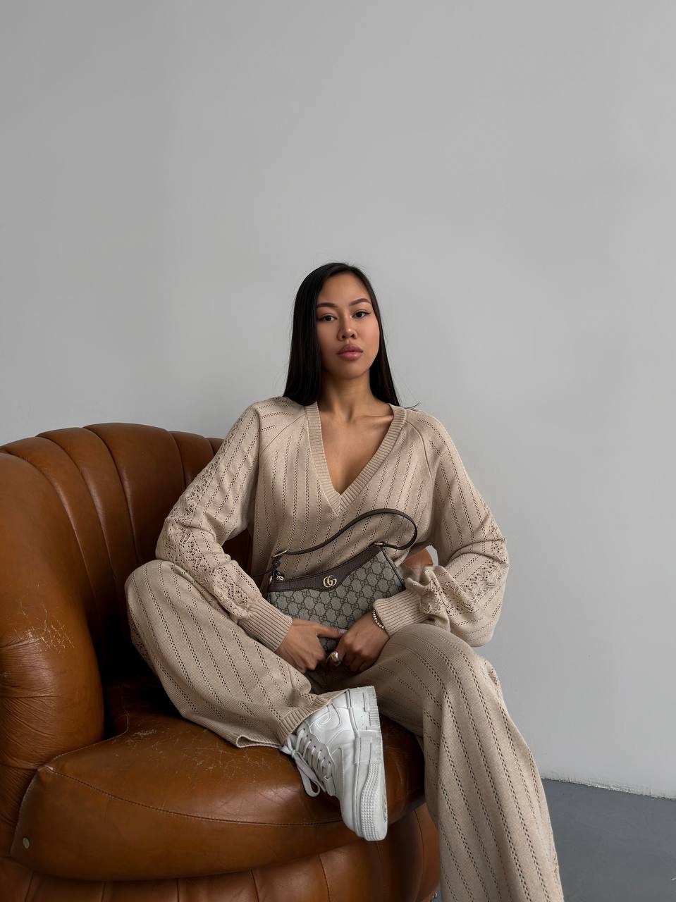 Woman in a beige outfit sitting on a brown leather couch holding a handbag.