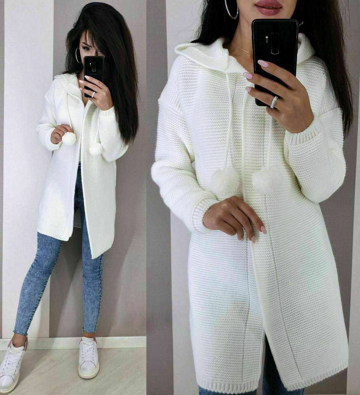 Woman wearing a white knit cardigan and blue jeans, taking a mirror selfie.