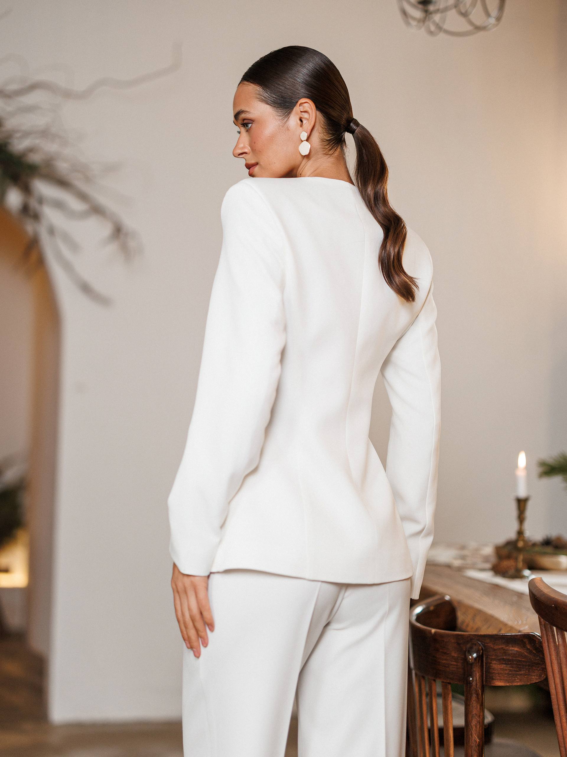 Woman in a white suit standing in a room with wooden furniture and a candle.