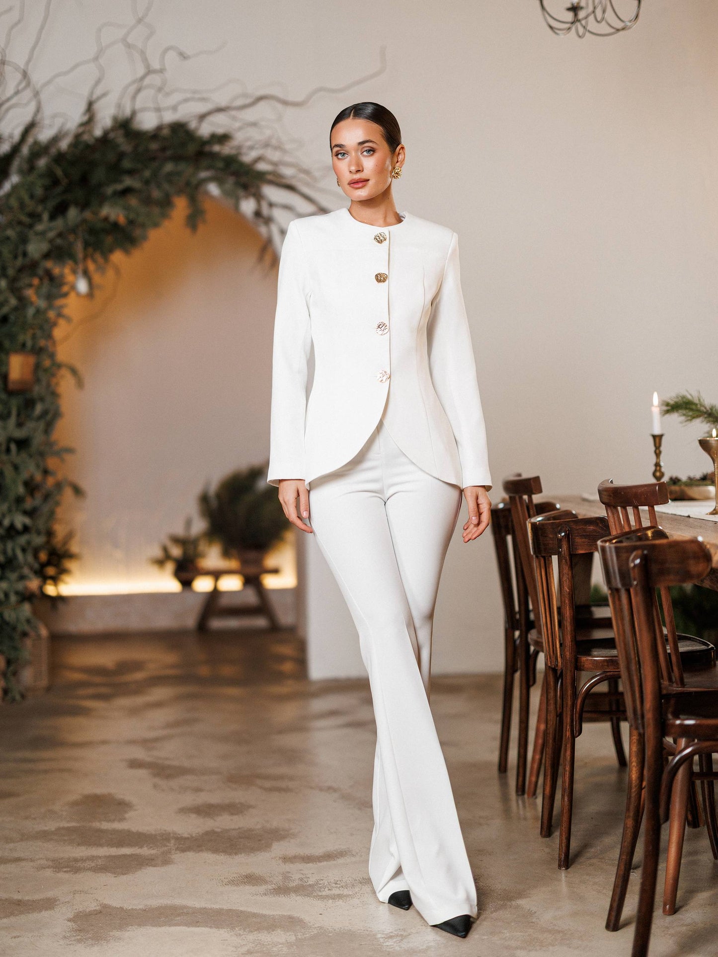 Woman in a white suit standing in a decorated room with wooden chairs and a table.
