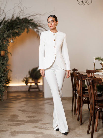 Woman in a white suit standing in a decorated room with wooden chairs and a table.