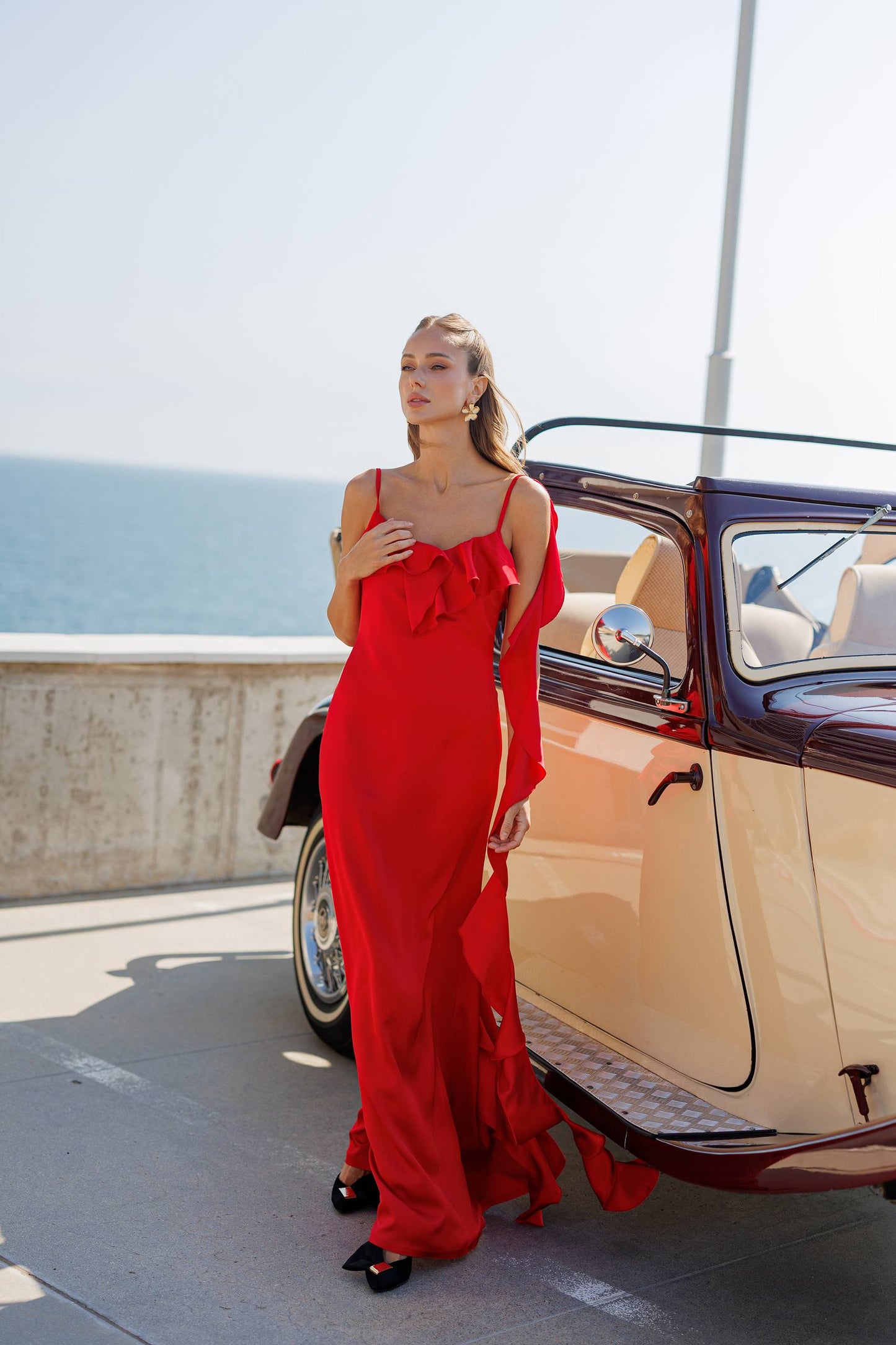 Woman in a red dress standing next to a vintage car by the sea.