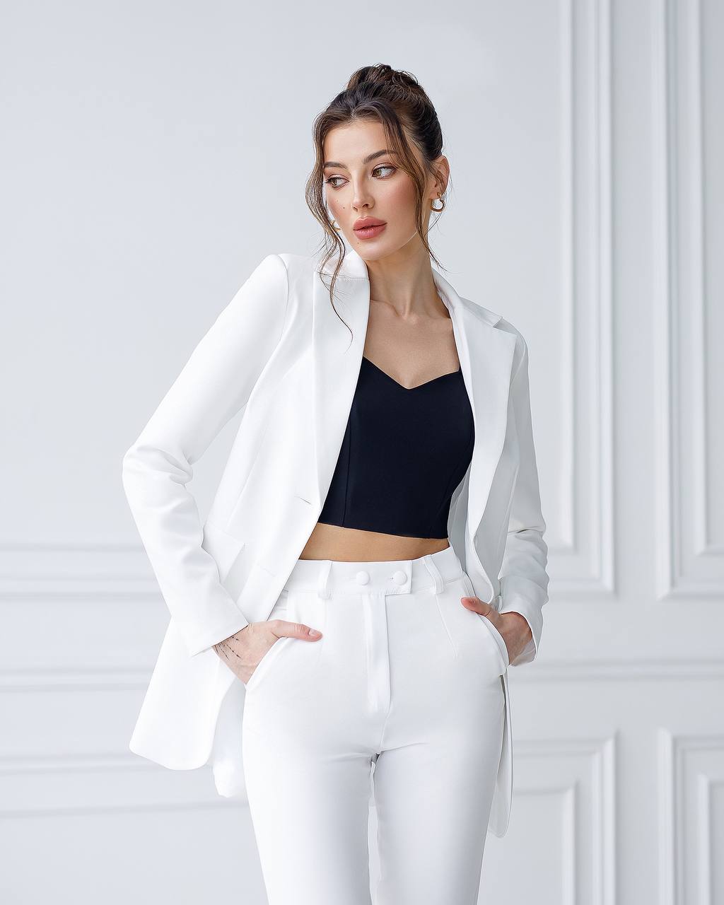 Woman wearing a white blazer and pants with a black top against a white wall.