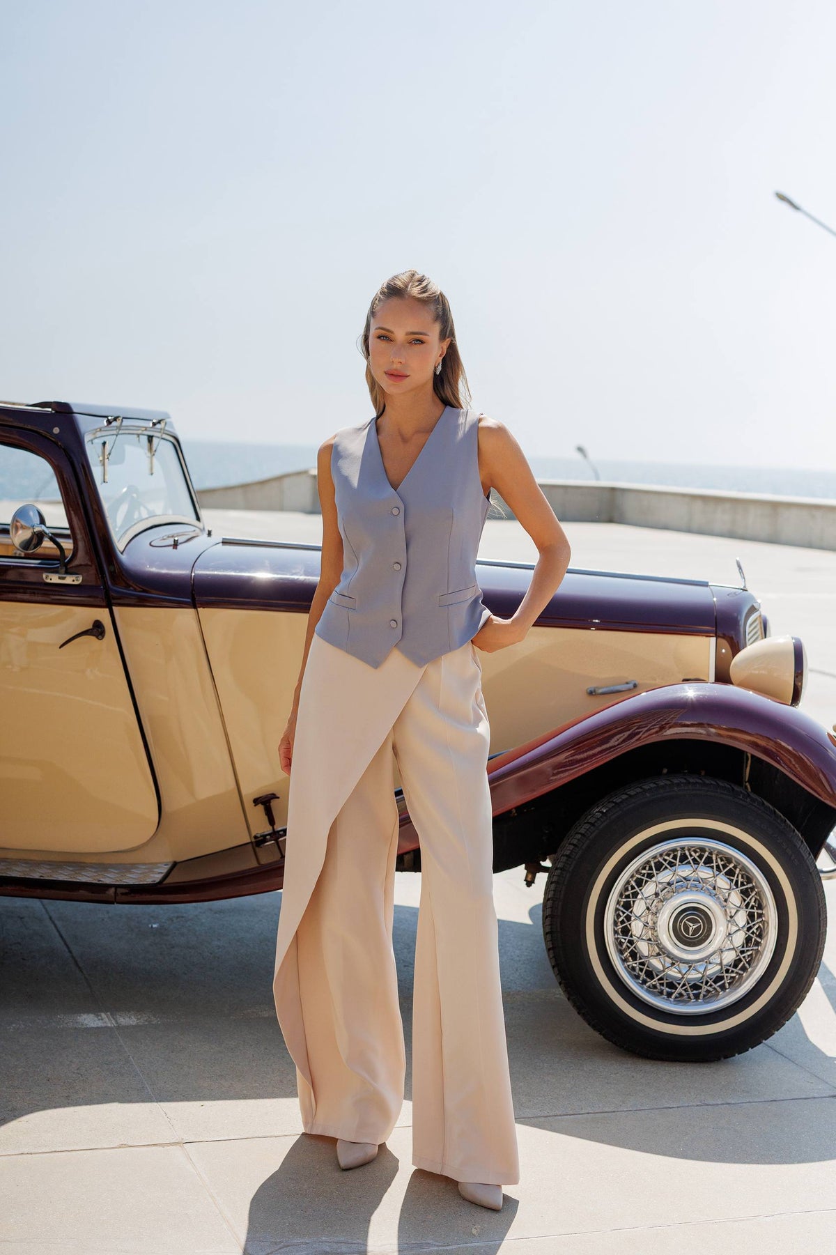 Woman in a sleeveless top and wide-leg pants standing next to an old-fashioned car on a sunny day.