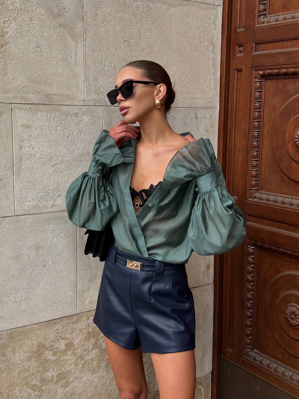 Woman wearing a green blouse and blue shorts standing against a stone wall.