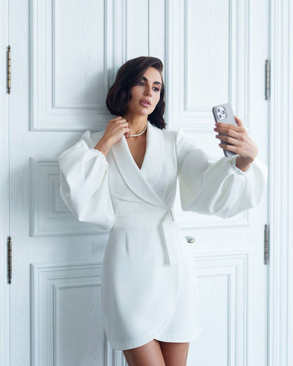 Woman in a white dress taking a selfie against a white paneled wall.