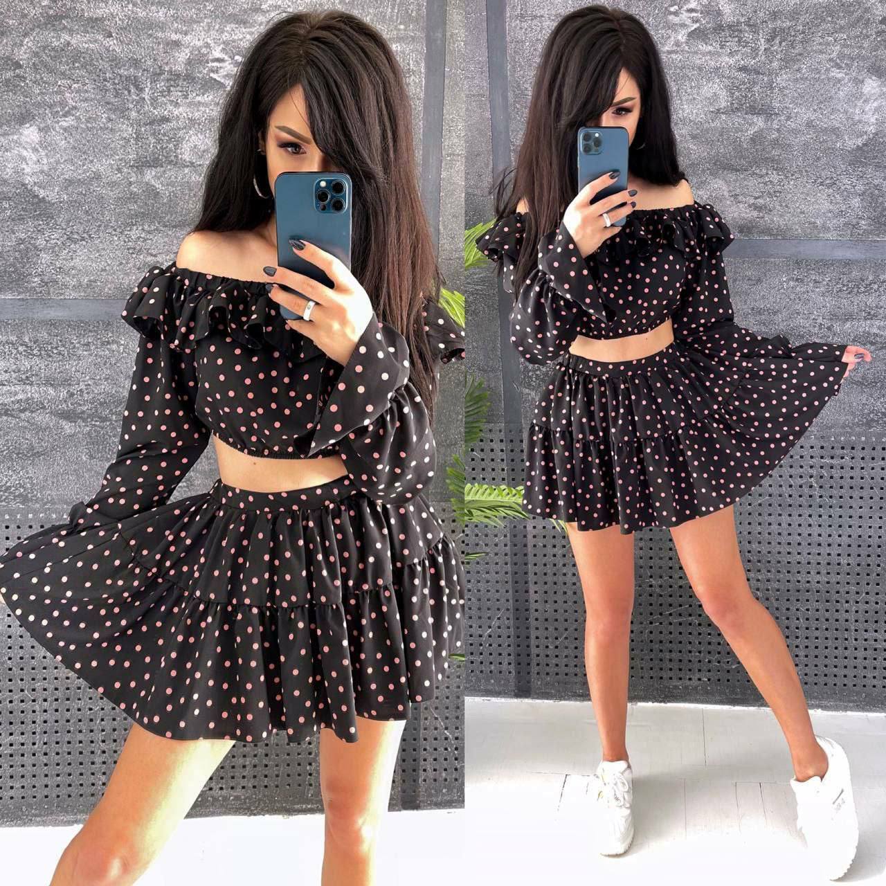 Woman in a black polka dot dress taking a mirror selfie against a gray textured wall.
