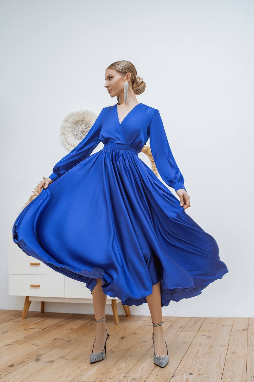 Woman wearing a blue dress with long sleeves in a room with wooden flooring and a white wall.