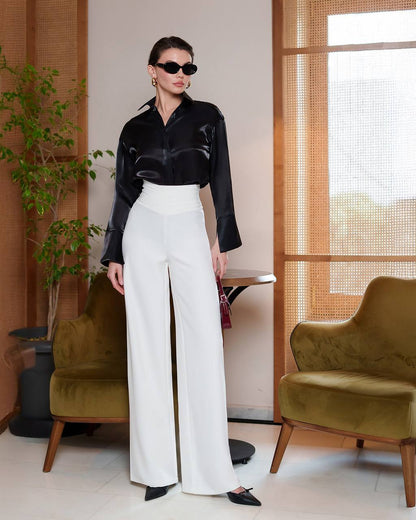 Woman in black blouse and white pants standing in a modern interior setting with green chairs and plants.